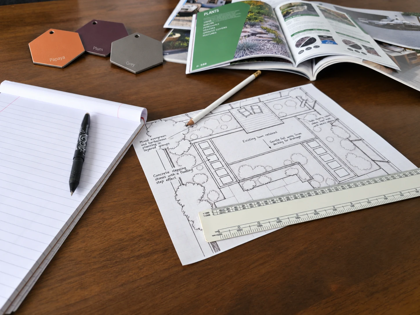 Garden design drawing with a pencil, ruler, blank notepad, planting book and papaya, plum and grey material samples