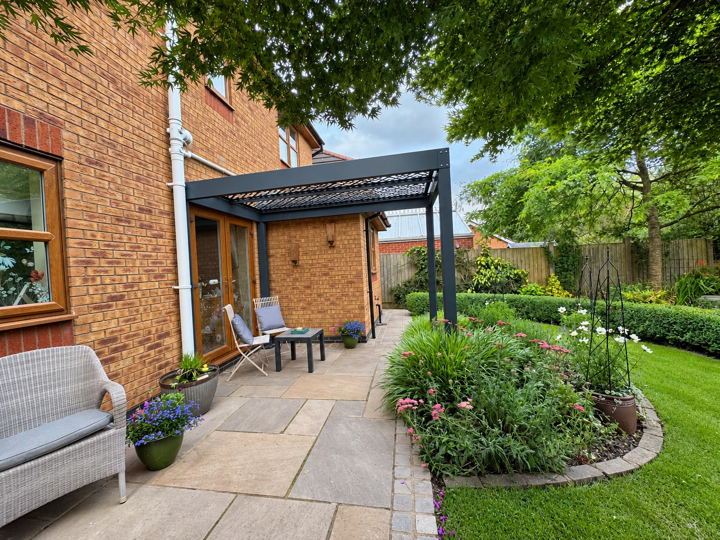 Grey pergola above a paved seating area beside colourful planting and lawn
